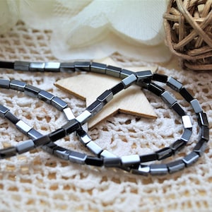 Peut inclure: Un brin de perles carrées noires et argentées. Les perles sont disposées en cercle et reposent sur une surface en crochet blanc.