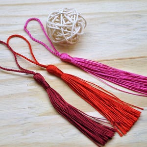 Puede incluir: Tres borlas decorativas en tonos burdeos intenso, rojo brillante y rosa vibrante, cada una con un cordón en bucle en la parte superior. Una pequeña bola esférica de ratán blanco trenzado se sitúa detrás de las borlas, añadiendo textura.