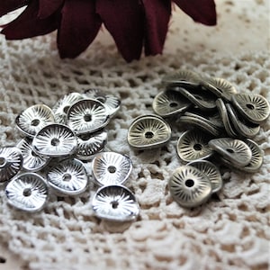 Op de afbeelding: Een verzameling zilverkleurige metalen kralenkapjes met een zonnestraalontwerp. De kralenkapjes zijn gerangschikt op een witte, open geweven stof, met een donkerrode bloem op de achtergrond.