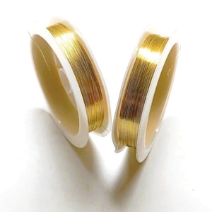 Può includere: Due bobine di filo color oro sono mostrate su sfondo bianco. Il filo è avvolto strettamente su bobine di plastica bianca. Il filo ha una finitura lucida e metallica, suggerendo che sia fatto di un metallo prezioso o di una lega dorata. Ideale per la creazione di gioielli.