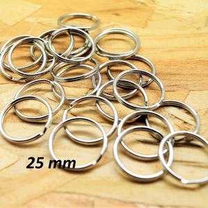 Puede incluir: Un conjunto de llaveros plateados, cada uno con un diseño circular. Los llaveros están dispersos sobre una superficie de madera. El texto "25 mm" es visible, indicando el diámetro de los anillos.