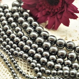 Peut inclure: Plusieurs rangées de perles polies gris foncé de différentes tailles. Une fleur bordeaux foncé est visible en arrière-plan. Les perles ont une surface réfléchissante, possiblement métallique ou en verre. Ces perles sont probablement pour la création de bijoux.