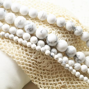 May include: Close-up of white marble beads with grey veining, in different sizes, arranged on a cream crocheted fabric. The beads are spherical and smooth, offering a natural and elegant appearance.