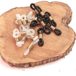 以下が含まれることがあります： ゴールドカラーの金属アクセントが付いた、黒とクリアのメガネホルダー。メガネホルダーは木製の表面に表示されています。ホルダーは、メガネを安全かつアクセスしやすくするために首の周りに着用するように設計されています。