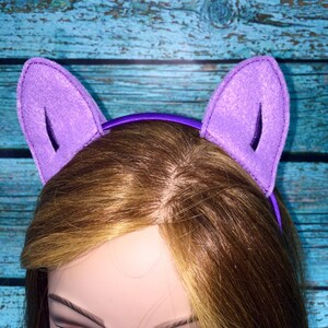 May include: A purple headband featuring two upright, triangular felt cat ears. The ears are constructed from a light purple felt material with a darker purple inner detail, attached to a smooth, solid purple plastic headband base.