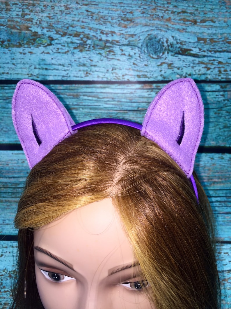 May include: A purple headband featuring two upright, triangular felt cat ears. The ears are constructed from a light purple felt material with a darker purple inner detail, attached to a smooth, solid purple plastic headband base.
