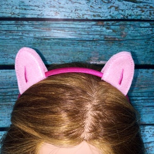 May include: A bright pink headband featuring two upright, soft felt cat ears in a lighter shade of pink. The headband has a smooth, shiny finish and the ears are constructed with a stitched border and a vertical seam detail on the front surface.
