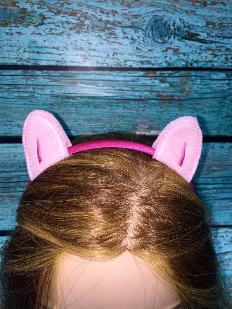 May include: A bright pink headband featuring two upright, soft felt cat ears in a lighter shade of pink. The headband has a smooth, shiny finish and the ears are constructed with a stitched border and a vertical seam detail on the front surface.