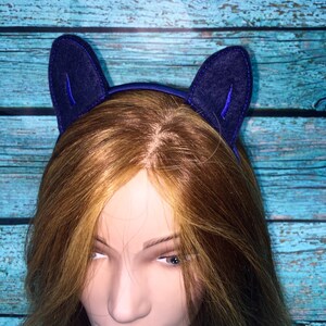 May include: A dark blue headband featuring two upright, pointed felt cat ears. The ears are constructed from a deep navy fabric with a stitched border and a smaller, vibrant blue inner ear detail. The headband is worn on a mannequin head.
