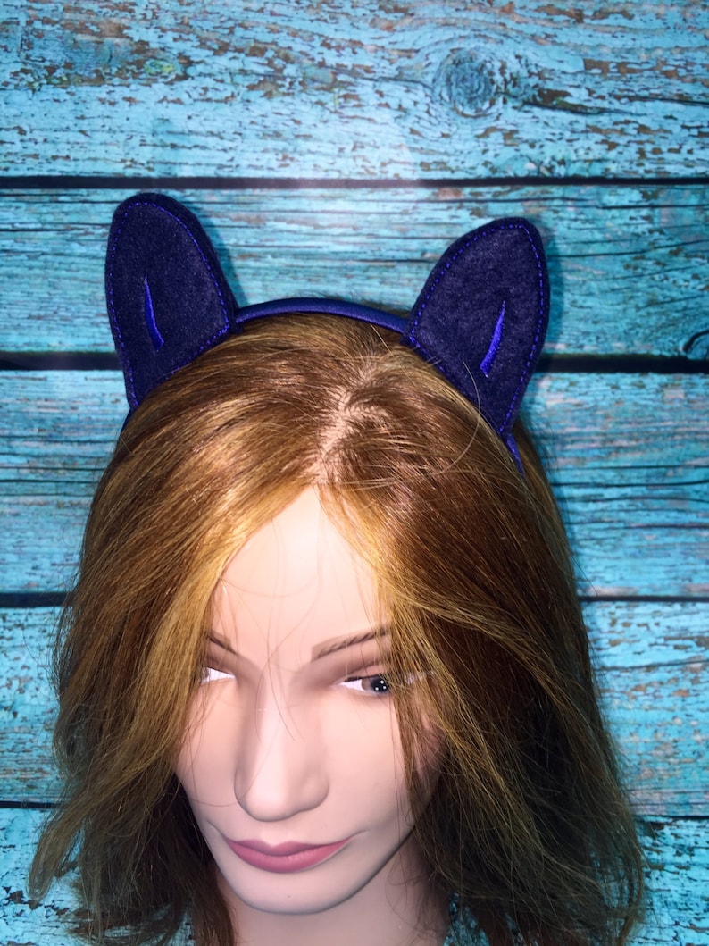 May include: A dark blue headband featuring two upright, pointed felt cat ears. The ears are constructed from a deep navy fabric with a stitched border and a smaller, vibrant blue inner ear detail. The headband is worn on a mannequin head.