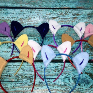 May include: A collection of cat ear headbands in various colors including purple, dark blue, white, pink, yellow, tan, and light blue. Each headband features two triangular fabric ears with contrasting inner ear details attached to a thin band.