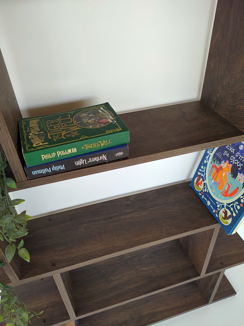 Pu&ograve; includere: Una libreria in legno marrone con tre ripiani. Il ripiano superiore ha due libri impilati uno sopra l'altro. Il libro superiore &egrave; verde con il titolo "The Tiger Rising" di Philip Pullman. Il libro inferiore &egrave; blu con il titolo "Northern Lights" di Philip Pullman. Il ripiano intermedio &egrave; vuoto. Il ripiano inferiore ha un libro per bambini con un'illustrazione colorata di animali su uno sfondo bianco. Il titolo del libro &egrave; "Eva o's Big Book of Bugs".