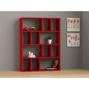 May include: A red bookshelf with 12 compartments, some of which are filled with books and a potted plant. The bookshelf is standing on a wooden floor in front of a gray wall.