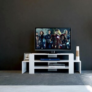May include: White TV stand with three shelves. The stand has a modern design and is made of wood. A flat screen TV is on top of the stand. The TV is playing a movie with a group of people in it.