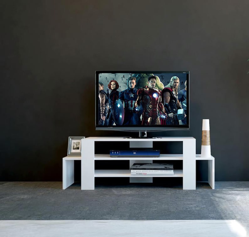 May include: White TV stand with three shelves. The stand has a modern design and is made of wood. A flat screen TV is on top of the stand. The TV is playing a movie with a group of people in it.