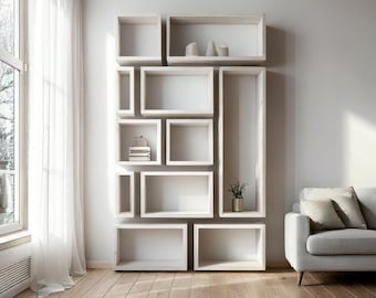 Op de afbeelding: Een moderne, witte wandplank met diverse rechthoekige en vierkante compartimenten. Sommige planken bevatten kleine decoratieve items. De unit staat tegen een lichtgrijze muur, met een houten vloer en een raam met transparante gordijnen.