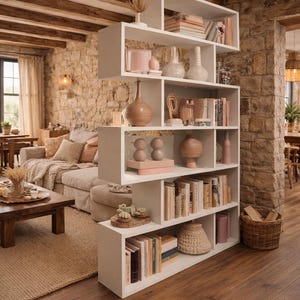 Op de afbeelding: Een witte, geometrische boekenkast gevuld met boeken, vazen en decoratieve objecten. De boekenkast dient als roomdivider en scheidt een woonkamer met een beige bank en een stenen muur. De kamer heeft een warme, uitnodigende sfeer.