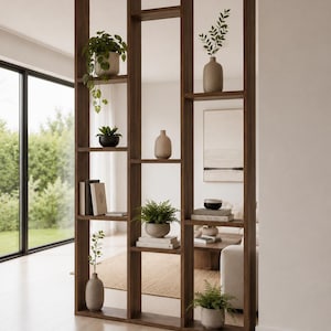 May include: A tall, dark brown wooden room divider featuring an open, asymmetrical grid design with multiple rectangular shelves. The shelves are decorated with various potted green plants, ceramic vases, and stacked books in a neutral colour palette.