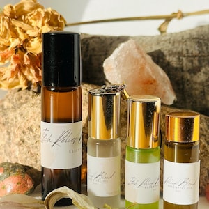 Puede incluir: Cuatro botellas de roll-on de aceite esencial con tapas doradas y etiquetas blancas. Las botellas son ámbar, transparentes y verdes. Las etiquetas dicen "Itch Relief Blend" y "Relax Blend Essential Oil". Flores secas y una roca en el fondo.