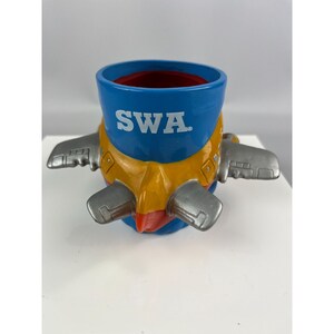 Puede incluir: Una taza de cer&aacute;mica azul y amarilla con forma de avi&oacute;n. La taza tiene las letras "SWA" impresas en el lateral. La taza tiene alas plateadas y un interior rojo.