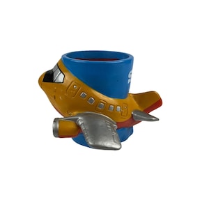 Puede incluir: Una maceta caprichosa con forma de avi&oacute;n en tonos amarillos, azules y rojos. La maceta presenta una gran abertura para plantas y detalles plateados en los motores. El dise&ntilde;o recuerda a un avi&oacute;n de dibujos animados.