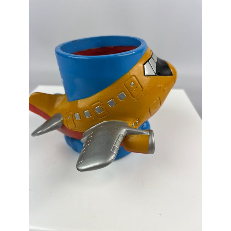 Puede incluir: Una maceta caprichosa con forma de avi&oacute;n. La maceta presenta un interior azul, un cuerpo amarillo con alas plateadas y un acento rojo en la cola. El dise&ntilde;o recuerda a un avi&oacute;n de dibujos animados.