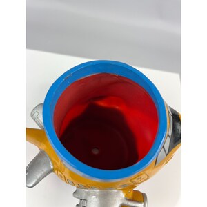 Puede incluir: Una maceta con forma de avi&oacute;n amarilla, azul, roja y plateada. La maceta tiene un borde azul, un interior rojo y alas plateadas. El exterior es amarillo con detalles en negro y plateado. La maceta est&aacute; dise&ntilde;ada para contener plantas.