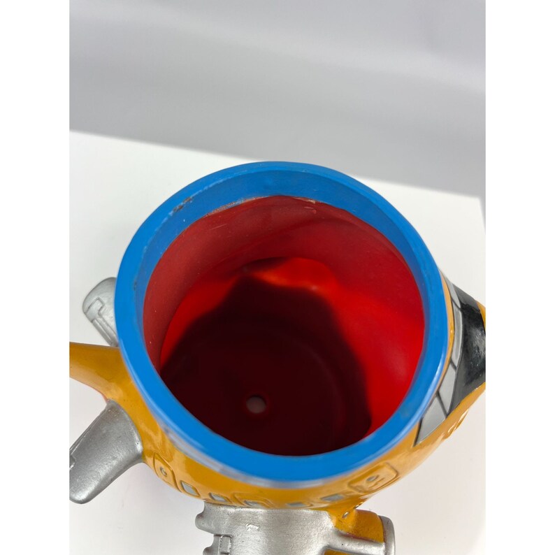 Puede incluir: Una maceta con forma de avi&oacute;n amarilla, azul, roja y plateada. La maceta tiene un borde azul, un interior rojo y alas plateadas. El exterior es amarillo con detalles en negro y plateado. La maceta est&aacute; dise&ntilde;ada para contener plantas.