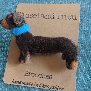 May include: A brown felt dachshund brooch with a blue felt collar. The brooch is on a brown card with the text "Tinsel and Tutu Brooches handmade in Shropshire".