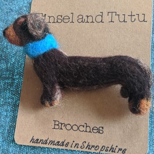 May include: A brown and black felt dachshund brooch wearing a blue felt scarf. The brooch is handmade in Shropshire and is labeled "Tinsel and Tutu Brooches".