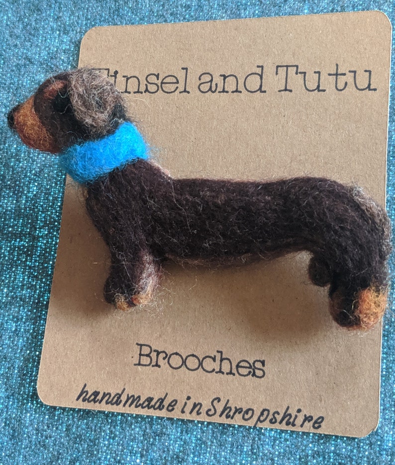 May include: A brown and black felt dachshund brooch wearing a blue felt scarf. The brooch is handmade in Shropshire and is labeled "Tinsel and Tutu Brooches".