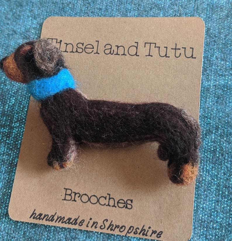 May include: A brown felt dachshund brooch with a blue felt collar. The brooch is on a brown card with the text "Tinsel and Tutu Brooches handmade in Shropshire".