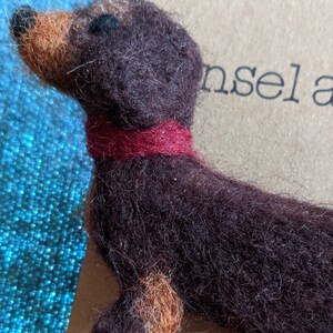 May include: A brown and tan felt dachshund figurine wearing a red felt collar. The figurine is sitting on a brown surface with the word "insel" printed on it.