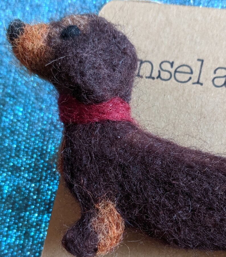 May include: A brown and tan felt dachshund figurine wearing a red felt collar. The figurine is sitting on a brown surface with the word "insel" printed on it.