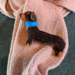 May include: A brown and black dachshund figurine wearing a blue collar, lying on a pink knitted scarf.