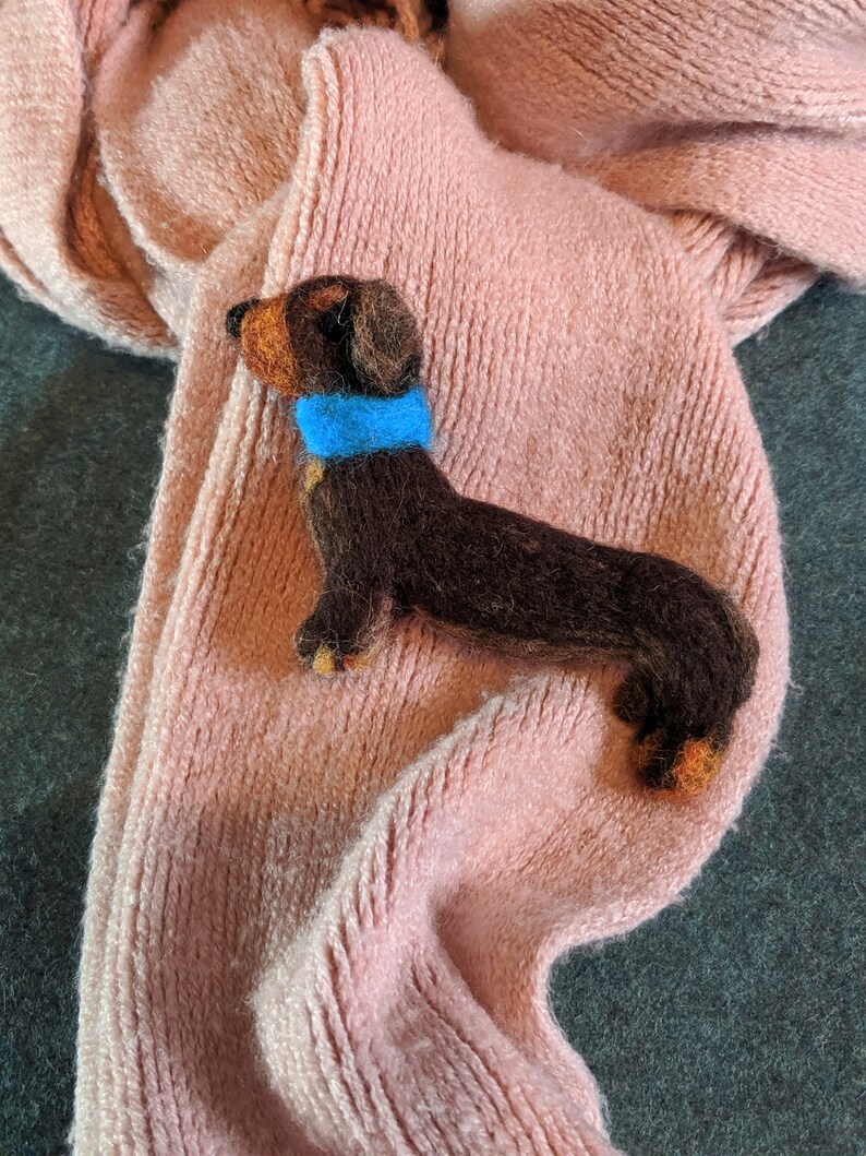 May include: A brown and black dachshund figurine wearing a blue collar, lying on a pink knitted scarf.