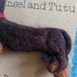 May include: A brown and black felt dachshund brooch with a red collar. The brooch is on a brown background with the text "Insel and Tutu Brooches" printed below.