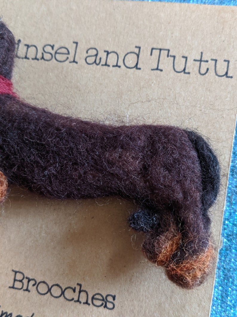 May include: A brown and black felt dachshund brooch with a red collar. The brooch is on a brown background with the text "Insel and Tutu Brooches" printed below.