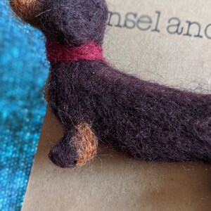 May include: A brown and tan felt dachshund brooch with a red felt collar. The brooch is on a brown paper card with the text "insel and" and "Brooch" printed on it.