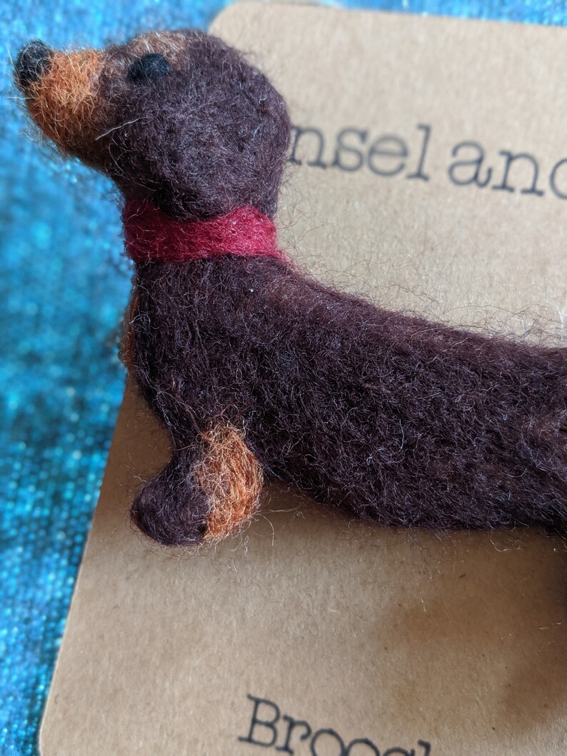 May include: A brown and tan felt dachshund brooch with a red felt collar. The brooch is on a brown paper card with the text "insel and" and "Brooch" printed on it.