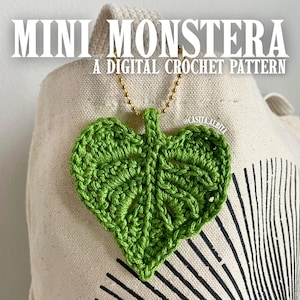 Op de afbeelding: Een heldergroene gehaakte bladhanger met een gouden ketting. Het blad is hartvormig met gedetailleerde steken. De afbeelding bevat de tekst "MINI MONSTERA A DIGITAL CROCHET PATTERN". De hanger hangt tegen een beige draagtas.