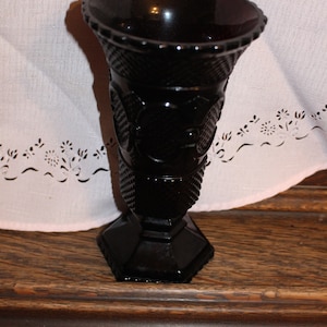 May include: A black glass vase with a textured design and a wide mouth. The vase has a square base and is decorated with raised floral patterns.