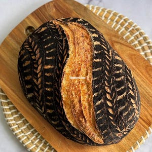 May include: An oval-shaped loaf of sourdough bread with a dark, charcoal-colored crust featuring intricate, light-colored wheat stalk patterns carved into the sides and a golden-brown, scored center ridge.
