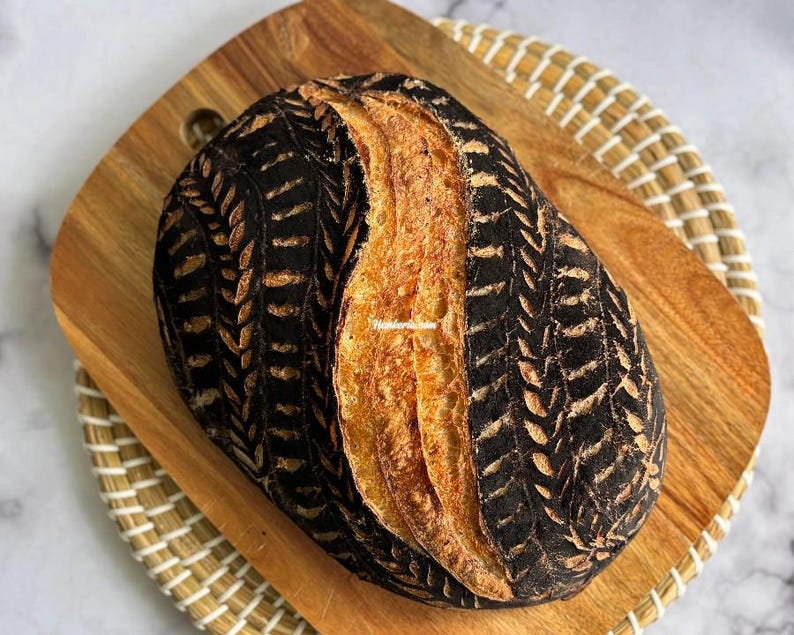 May include: An oval-shaped loaf of sourdough bread with a dark, charcoal-colored crust featuring intricate, light-colored wheat stalk patterns carved into the sides and a golden-brown, scored center ridge.