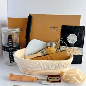 May include: A sourdough bread baking kit featuring a glass jar with a temperature band, a proofing basket with a liner, a wooden-handled scoring lame with razor blades, a pastry brush, a metal whisk, a dough scraper, and a black pouch.