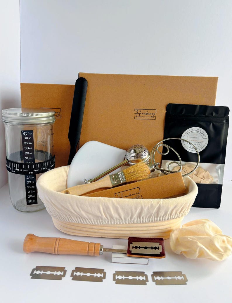 May include: A sourdough bread baking kit featuring a glass jar with a temperature band, a proofing basket with a liner, a wooden-handled scoring lame with razor blades, a pastry brush, a metal whisk, a dough scraper, and a black pouch.