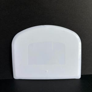 May include: A white plastic bowl scraper with a curved top edge and a straight bottom edge. The surface of the tool features a fine, textured grid pattern across the body, with a smooth, clear rectangular section in the center.