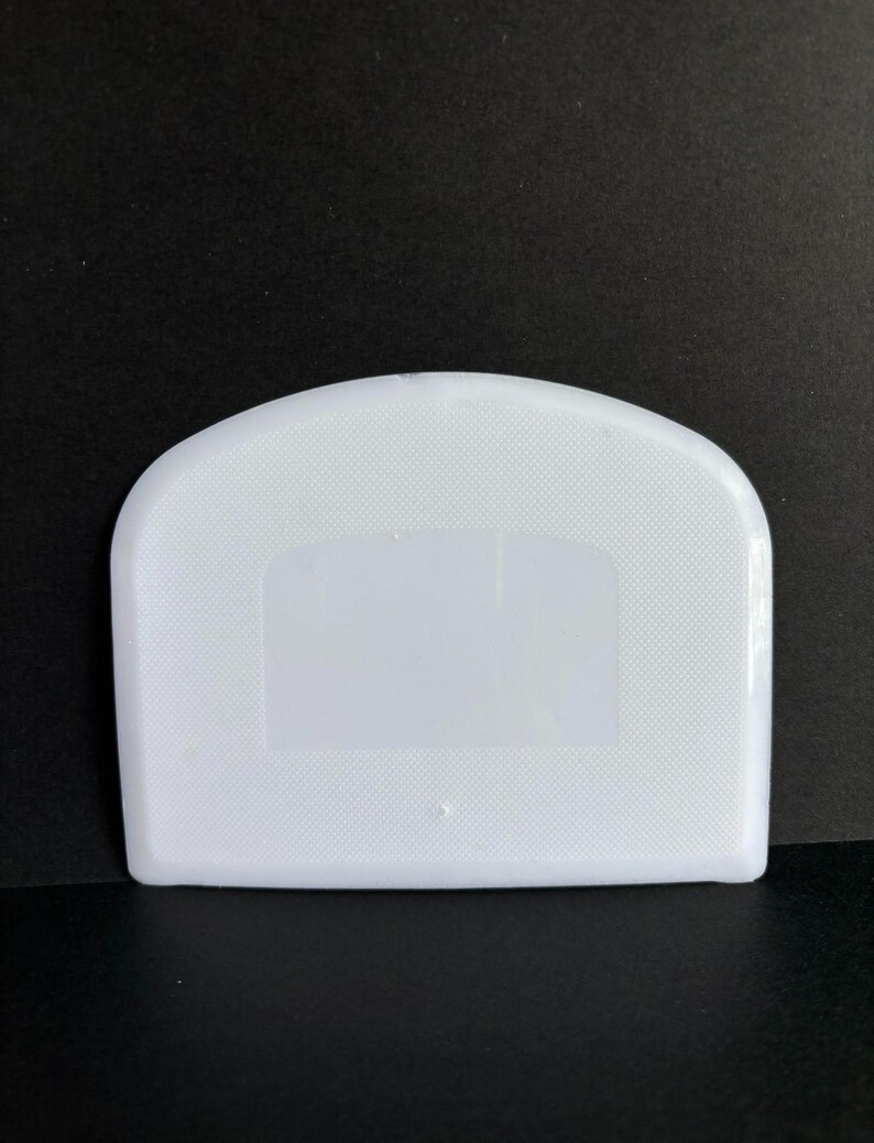 May include: A white plastic bowl scraper with a curved top edge and a straight bottom edge. The surface of the tool features a fine, textured grid pattern across the body, with a smooth, clear rectangular section in the center.