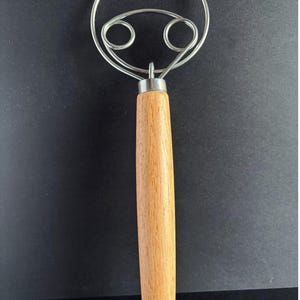 May include: A dough whisk featuring a long, cylindrical light brown wooden handle and a stainless steel wire head. The wire head consists of a large outer circular loop with two smaller, symmetrical inner loops connected to the handle.