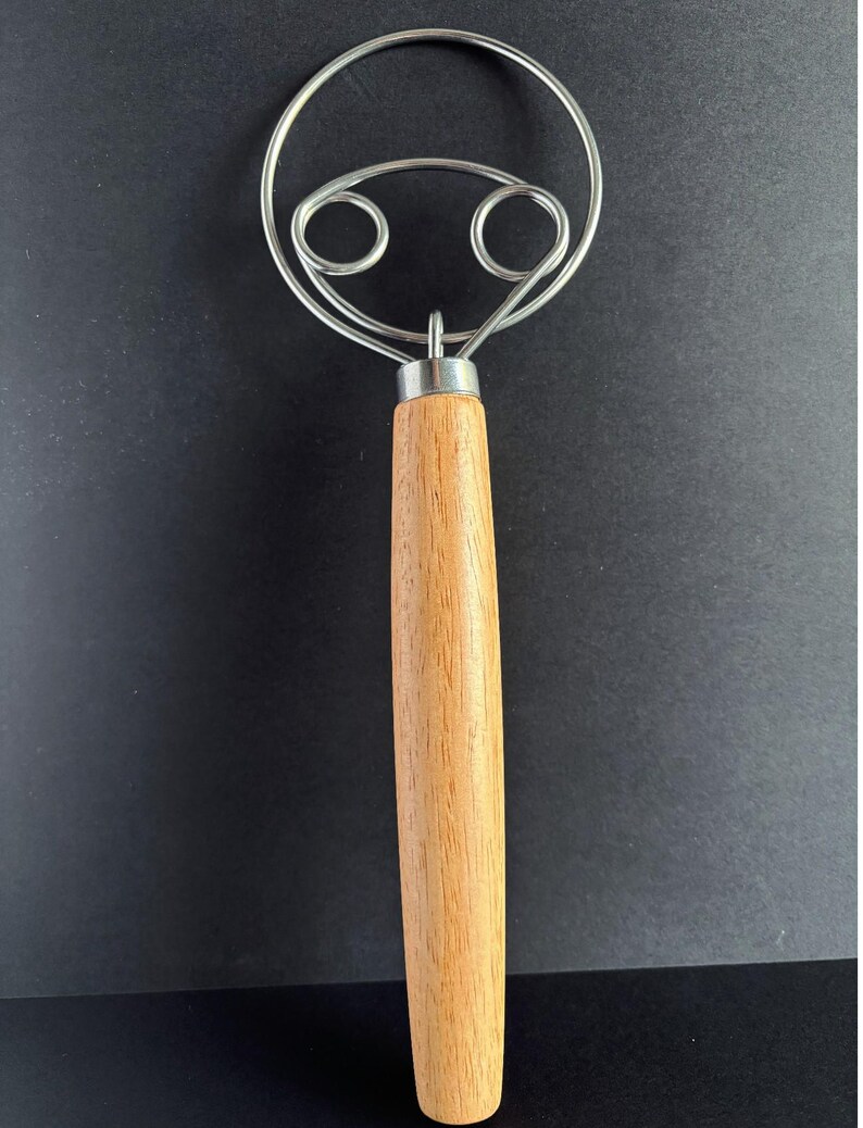 May include: A dough whisk featuring a long, cylindrical light brown wooden handle and a stainless steel wire head. The wire head consists of a large outer circular loop with two smaller, symmetrical inner loops connected to the handle.